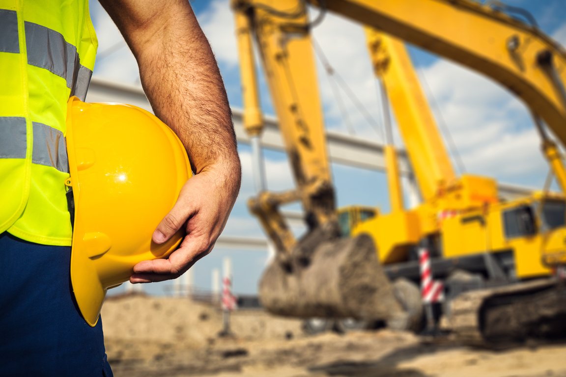 Construction worker holding a hard hat with excavators in the background